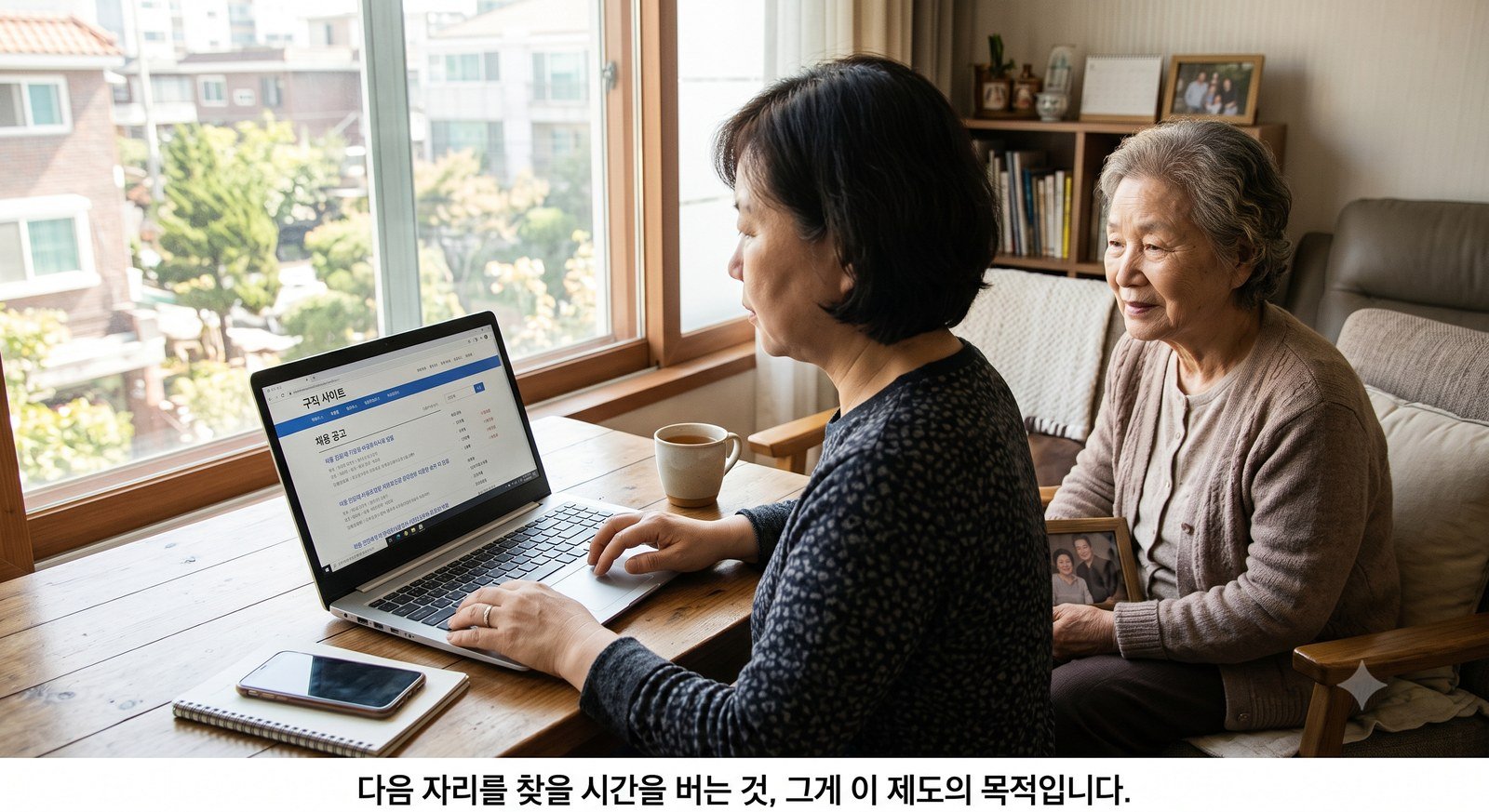 창가 식탁에서 노트북으로 구직 사이트를 보는 성인 자녀와 옆에 앉아 지켜보는 노인 어머니 — 다음 자리를 찾을 시간을 버는 것이 이 제도의 목적입니다